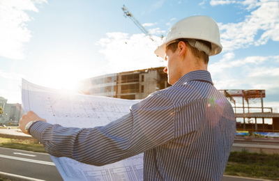 Architect in helmet with blueprints looks at camera in a building site Structural Engineering Services - Feasibility Study and Construction Inspection